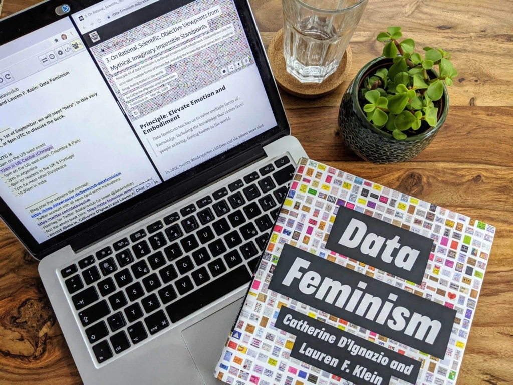 Laptop on a desk with the book "Data Feminism" on top and a little plant next to it. 